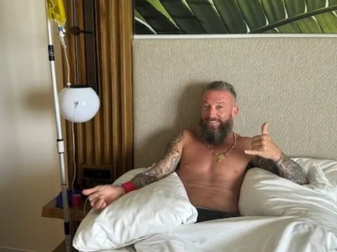 Man with tattoos in bed, making gesture.