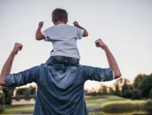 Child on shoulders, outdoors, flexing arms.